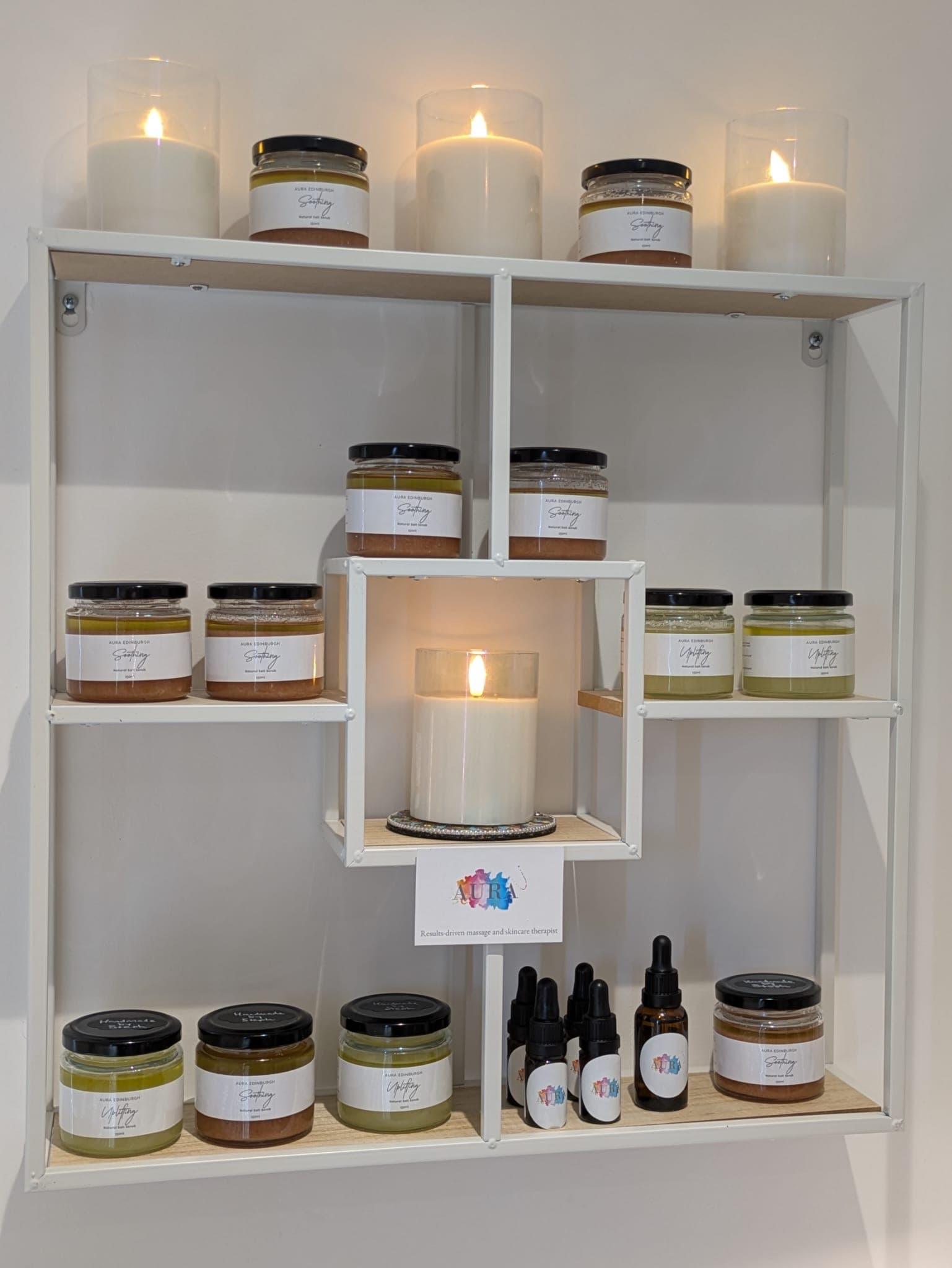 Aura's own labelled scrub jars and dropper bottles displayed on white shelves with lit candles