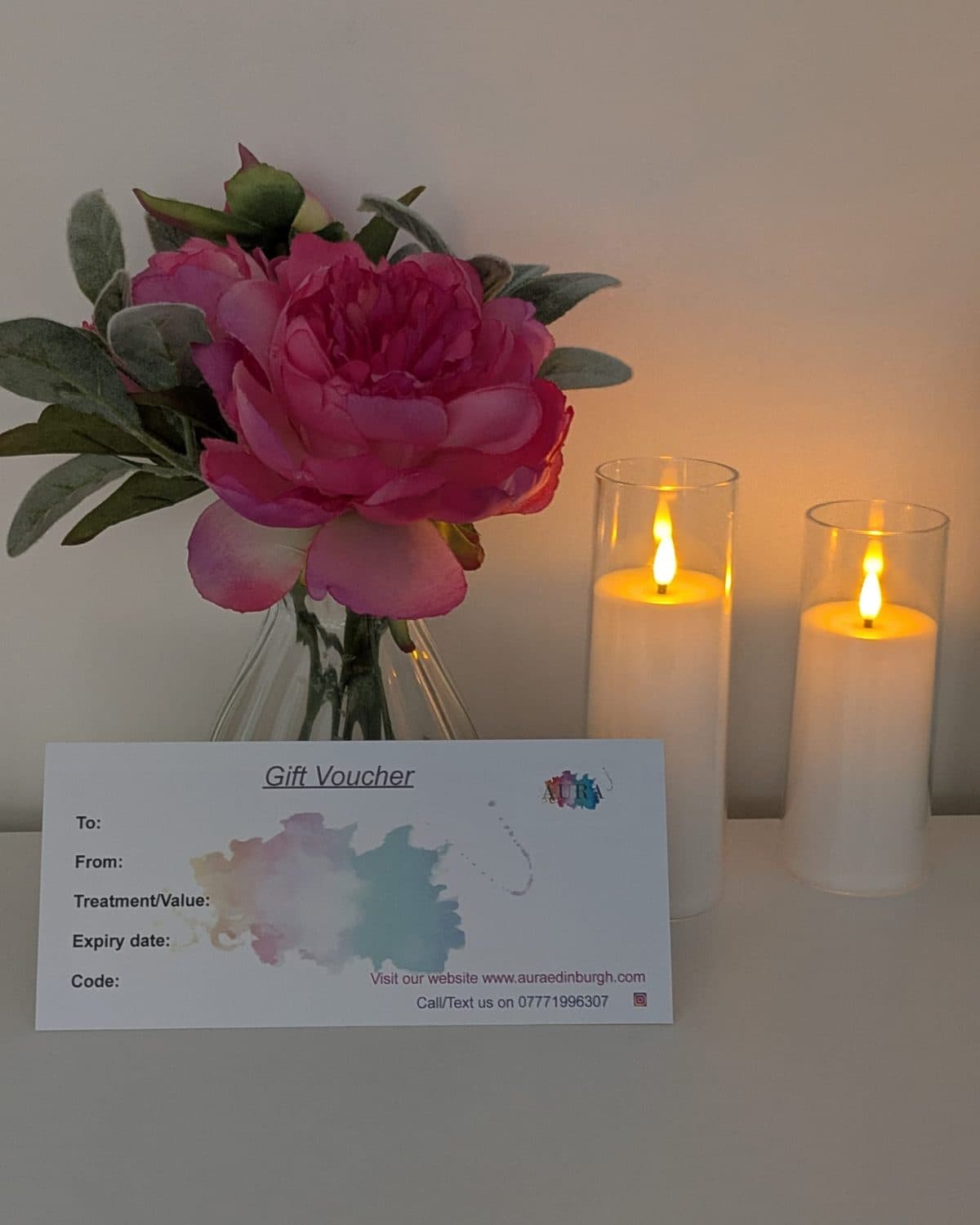 A printed Aura gift voucher beside pink peonies and two lit candles